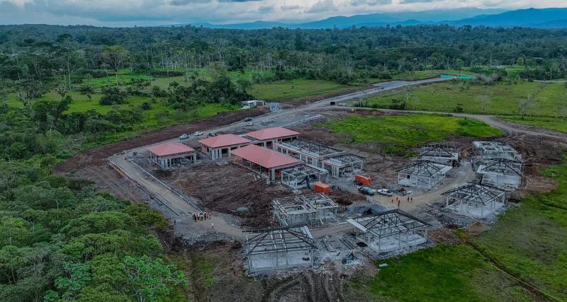Imagen aérea de un conjunto de edificaciones en construcción en una zona rural amazónica, rodeada de vegetación densa y montañas al fondo. Se observan varias estructuras con techos rojos y otras en fase inicial, además de maquinaria y trabajadores en el área.