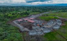 Imagen aérea de un conjunto de edificaciones en construcción en una zona rural amazónica, rodeada de vegetación densa y montañas al fondo. Se observan varias estructuras con techos rojos y otras en fase inicial, además de maquinaria y trabajadores en el área.